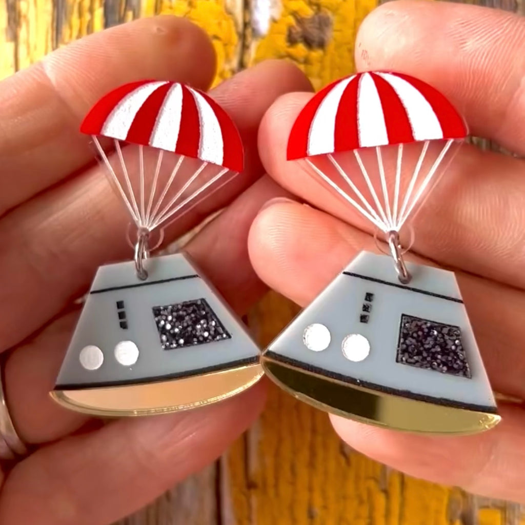 Space capsule earrings, with and red parachute topper and a grey capsule dangling underneath. Laser cut from acrylic. Earrings are being held up for a closeup view. 