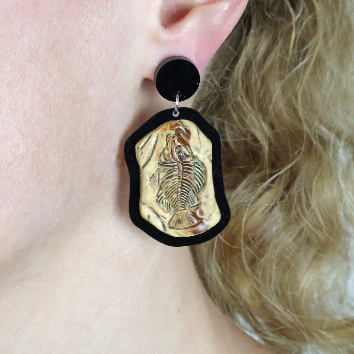 A pair of fish fossil themed acrylic and resin dangle earrings being modelled on an ear.