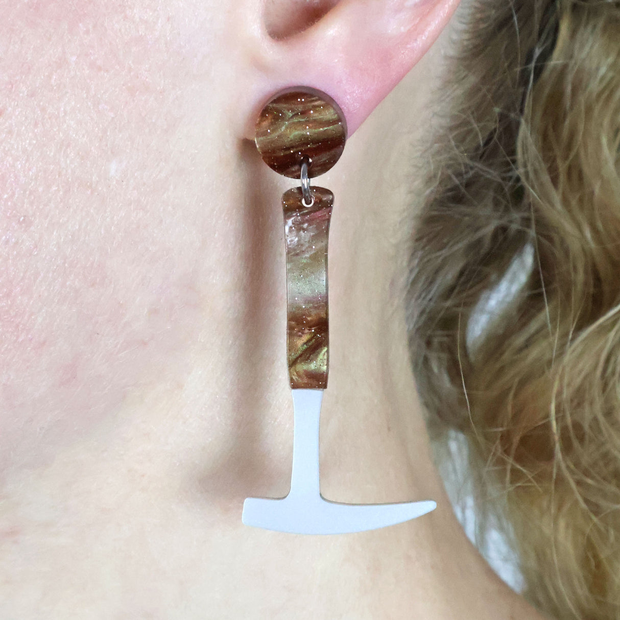 Laser cut acrylic fossil hammer shaped earrings being modelled on the ear of a blonde haired woman. The handle and earring topper has brown swirls and glitter.
