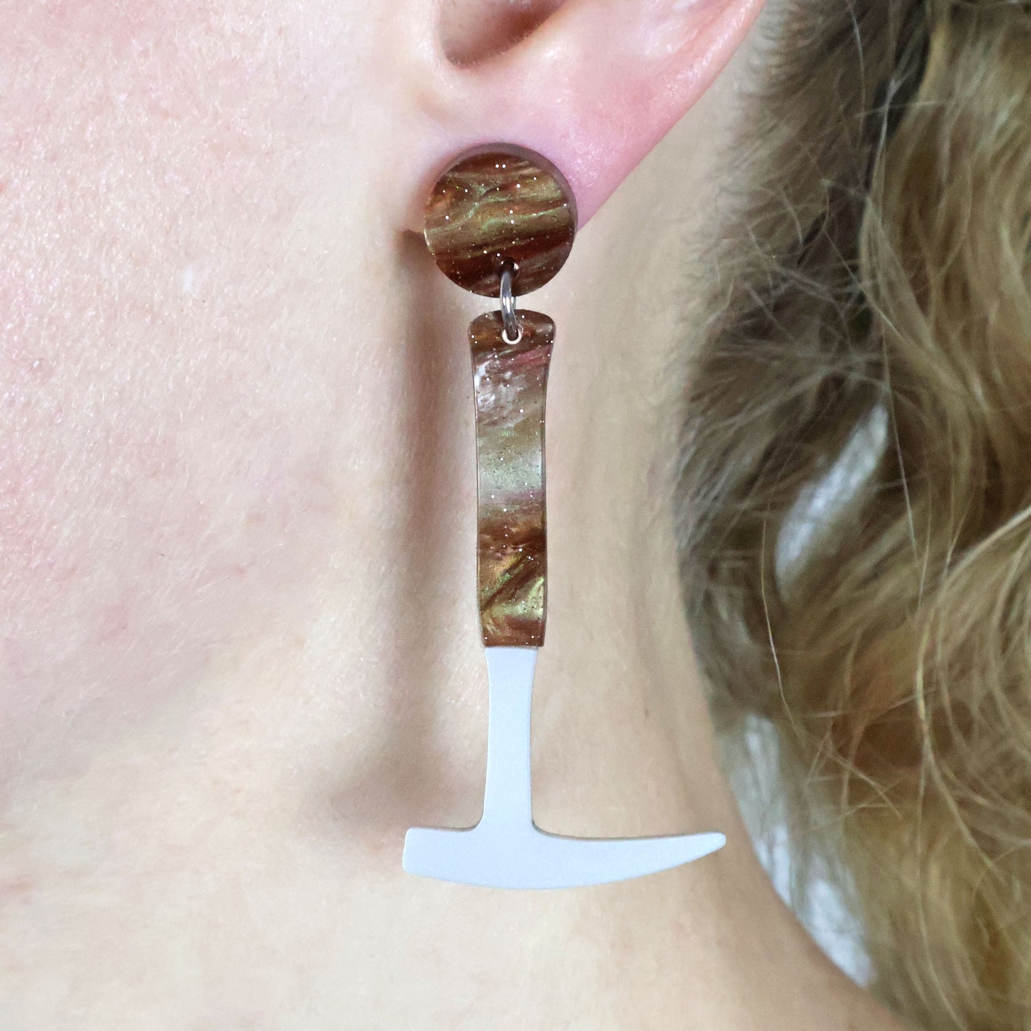 Laser cut acrylic fossil hammer shaped earrings being modelled on the ear of a blonde haired woman. The handle and earring topper has brown swirls and glitter.