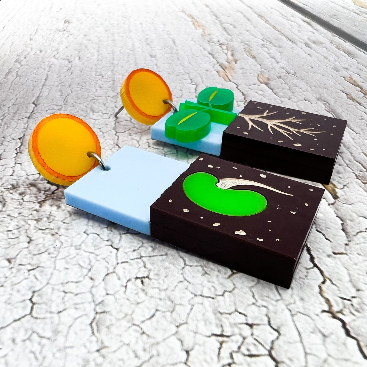 An angled view of a pair of asymmetrical earrings showing two stages in the germination process. One side shows a seed in the ground while the other side shows a seedling with roots. They hang form yellow 'sun' earring toppers. Laser cut and handpainted from acrylic.