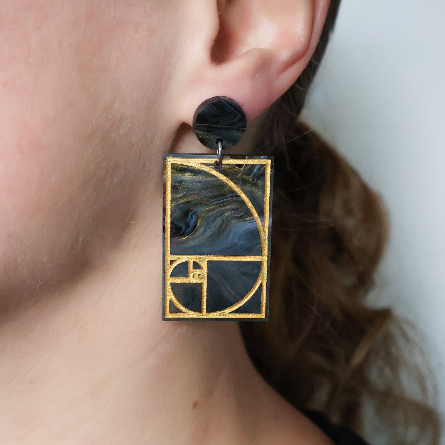 A pair of rectangular acrylic earrings displaying a gold painted golden ratio design in dark grey and gold acrylic, with a matching round acrylic topper, being modelled in an ear.