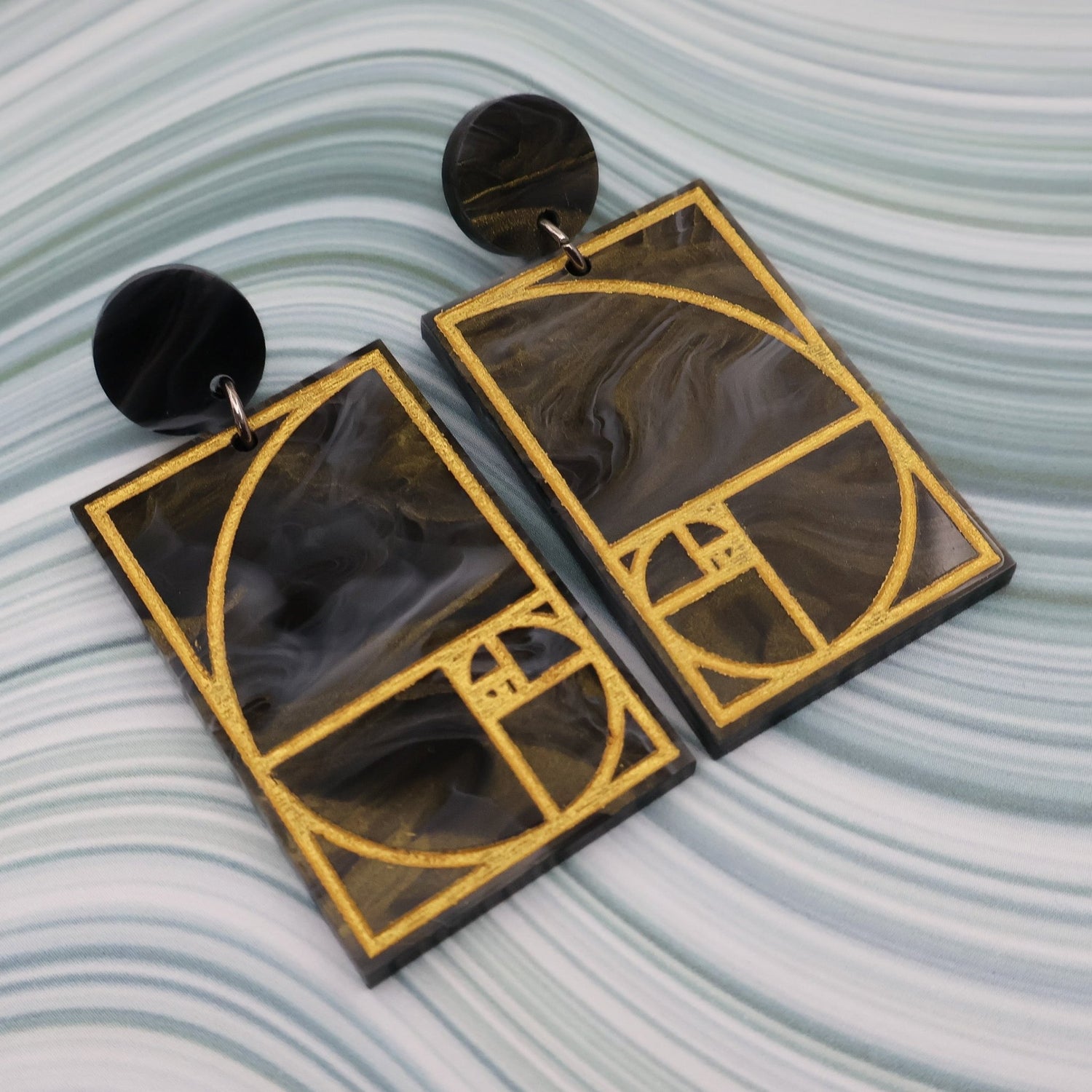 A pair of rectangular acrylic earrings displaying a gold painted golden ratio design in dark grey and gold acrylic, with a matching round acrylic topper, closeup view