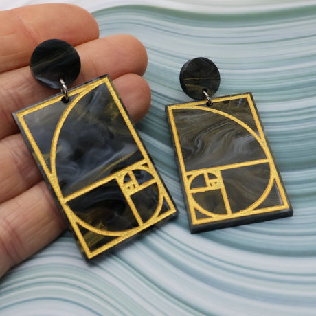 A pair of rectangular acrylic earrings displaying a gold painted golden ratio design in dark grey and gold acrylic, with a matching round acrylic topper, being held up by a hand