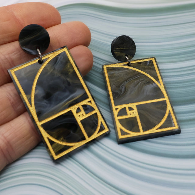 A pair of rectangular acrylic earrings displaying a gold painted golden ratio design in dark grey and gold acrylic, with a matching round acrylic topper, being held up by a hand