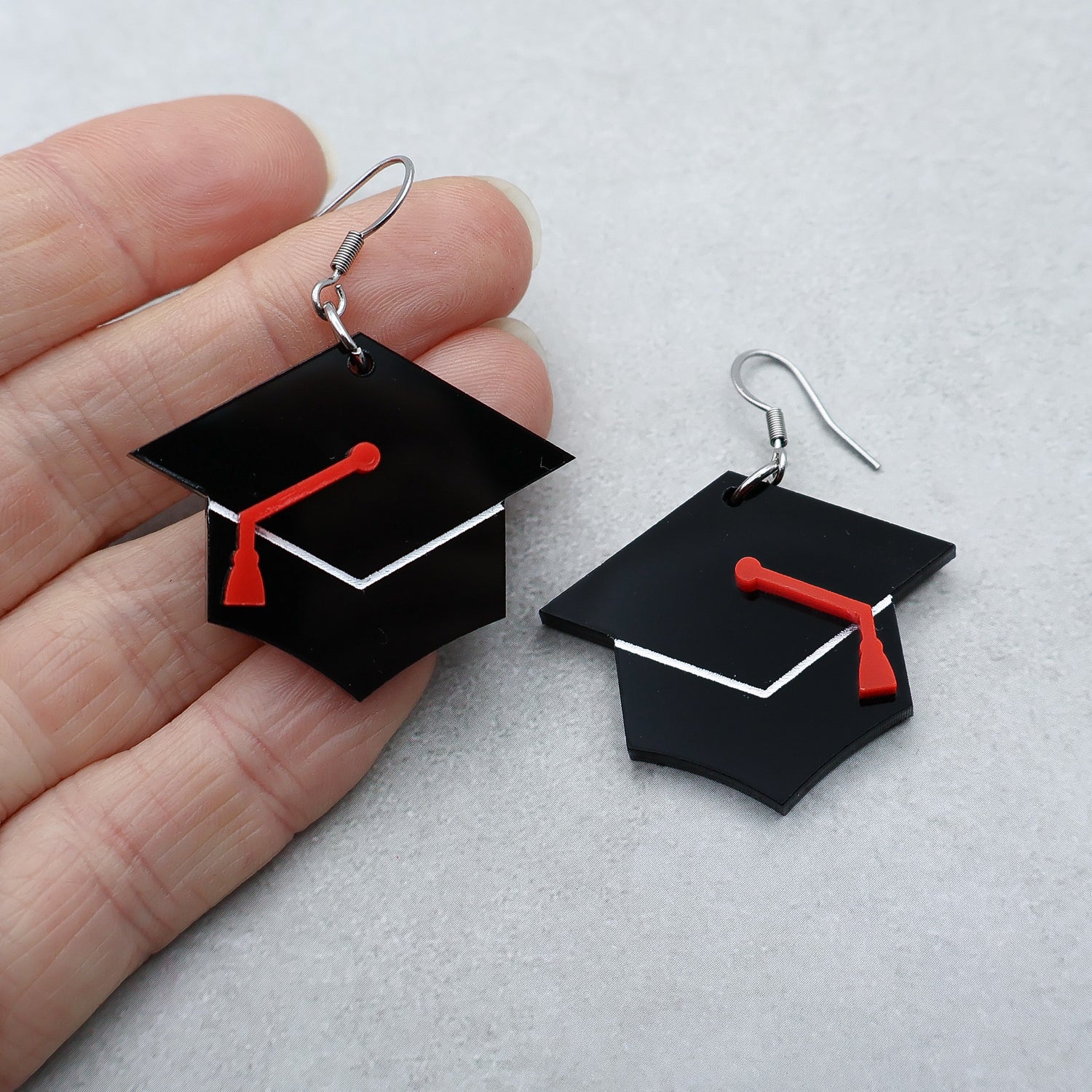 Closeup of graduation cap earrings with red tassles being held up in a hand. Laser cut acrylic caps hanging from stainless steel french hooks.