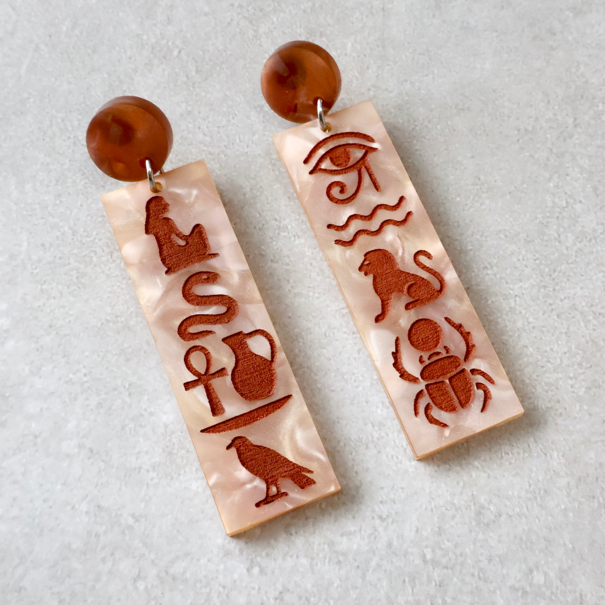 Pair of earrings with Egyptian hieroglyphic symbol designs held in a hand against a blue background.