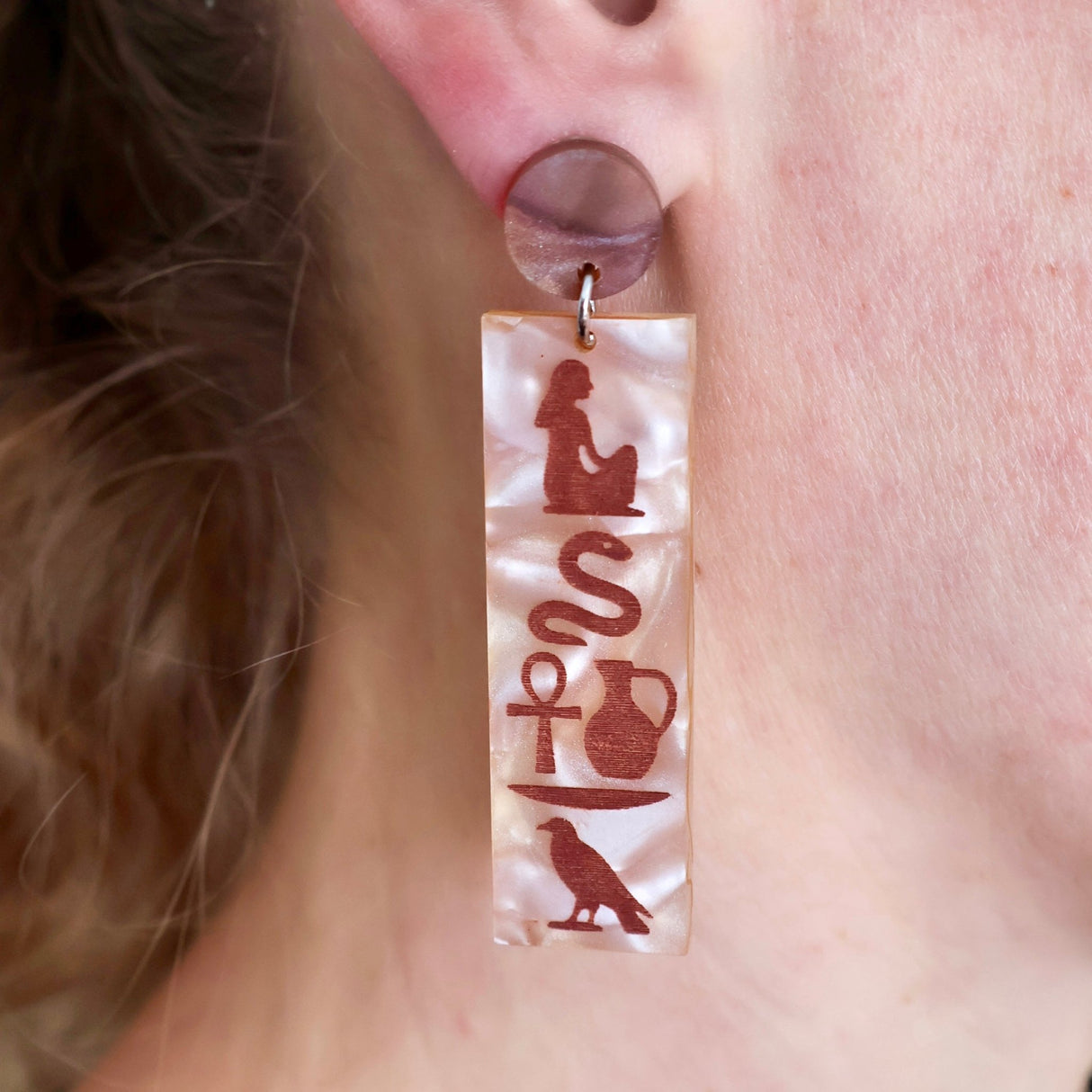 Pair of acrylic earrings with Egyptian hieroglyphic symbol designs  worn on a person's ear.