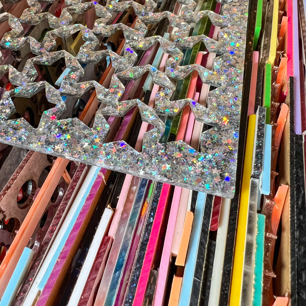 A stack of scrap acrylic sheets in various colours waiting to be recycled. The silver glitter sheet on top has had stars previously cut out of it. 