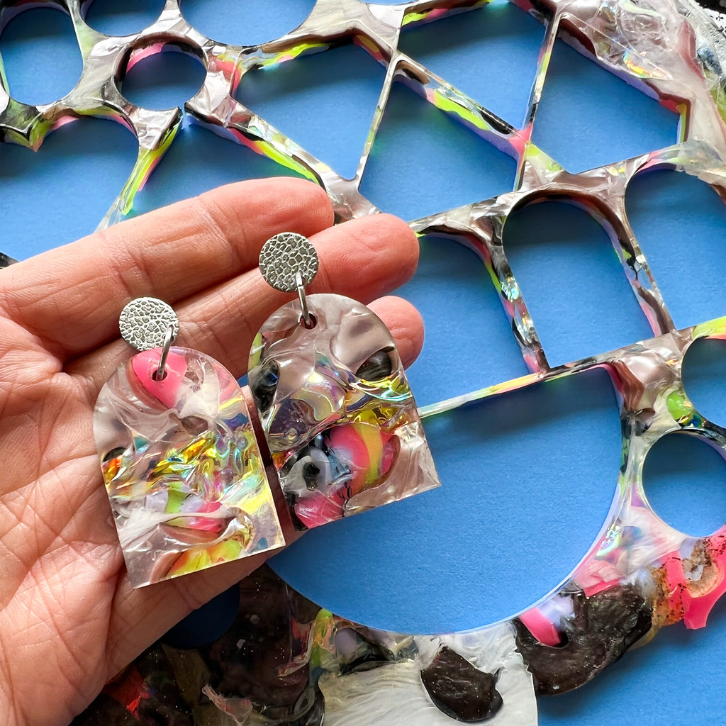 Some recycled arch shaped earrings being held above the sheet from which they were cut. The holes that were left behind in the sheet are evident.