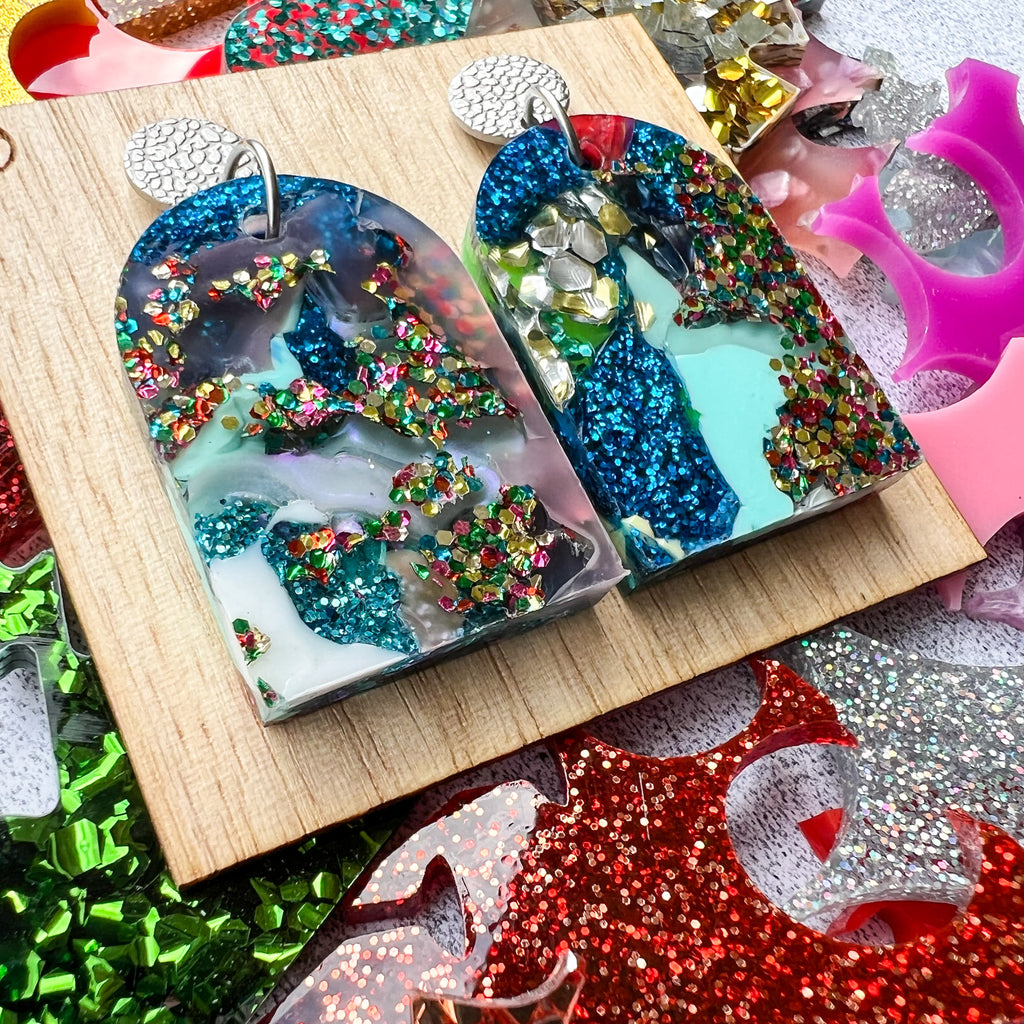 A pair of arch shaped earrings that were made in house from melted together acrylic scrap, and surrounded by pieces of colourful unprocessed scrap. 