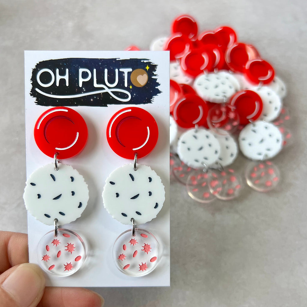 Dangle earrings comprising of a red blood cell topper, with a white blood cell and platelets dangling underneath. Laser cut and handpainted from acrylic. Earrings are being displayed on an Oh Pluto earring card. 