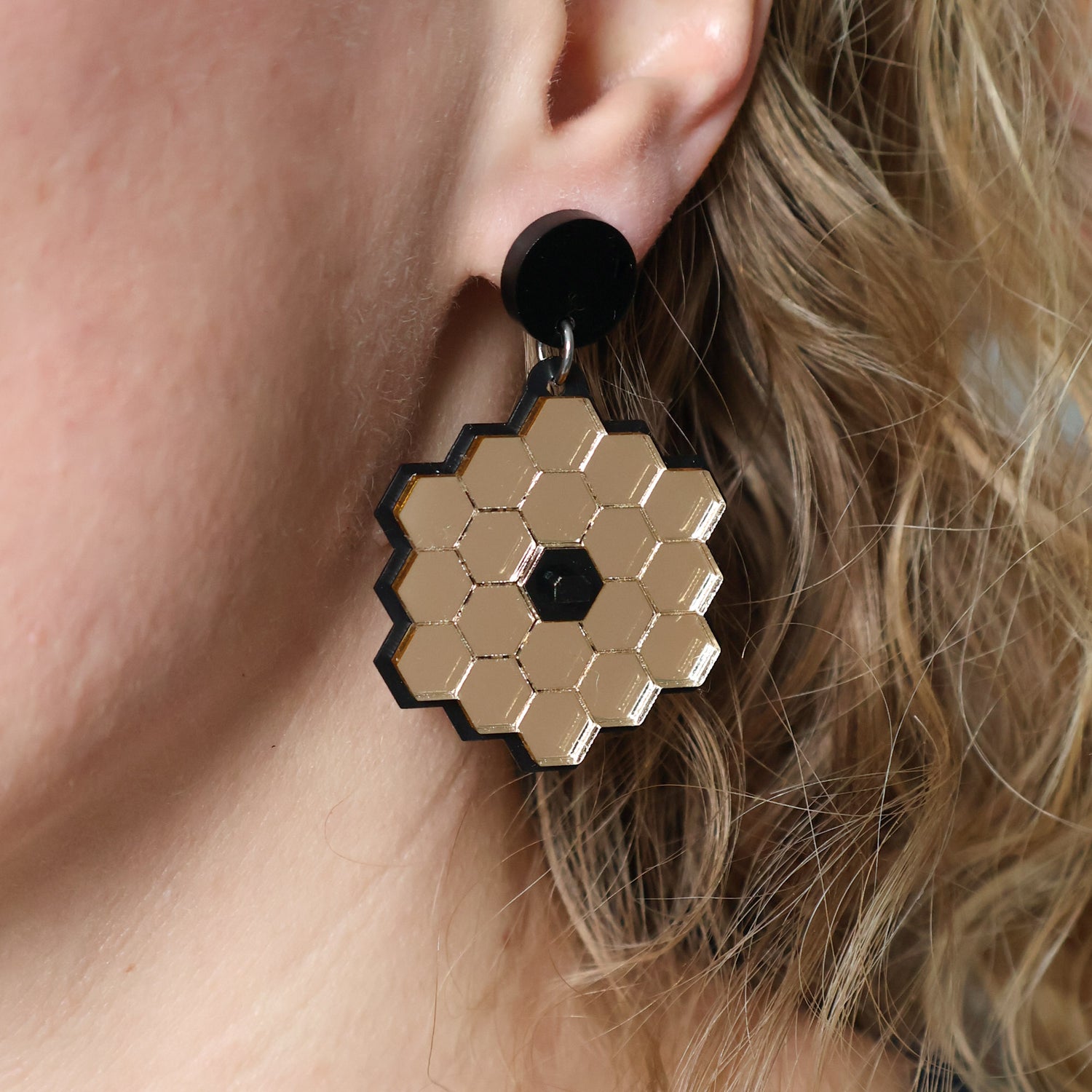 James Webb Space Telescope Earrings constructed from gold mirror acrylic and black acrylics, being modelled.