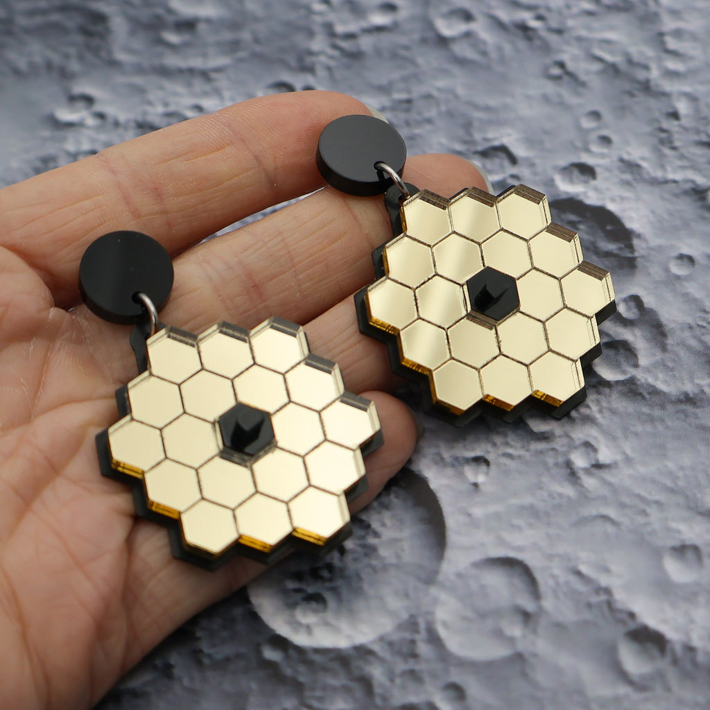 James Webb Space Telescope Earrings constructed from gold mirror acrylic and black acrylics, being held up, and showing the reflective properly of the mirror acrylic. 
