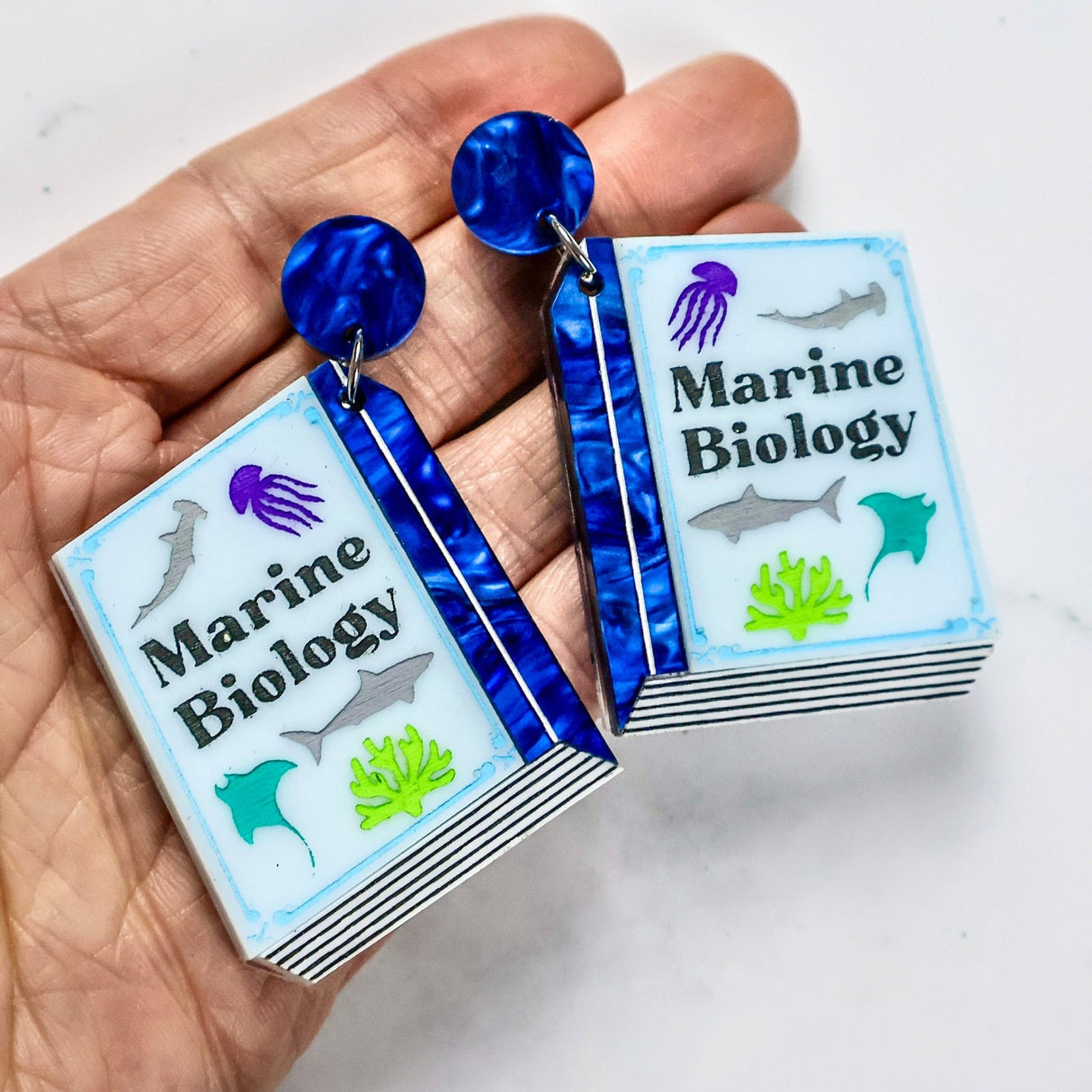 Earrings shaped like miniature books titled 'Marine Biology' held in a hand.