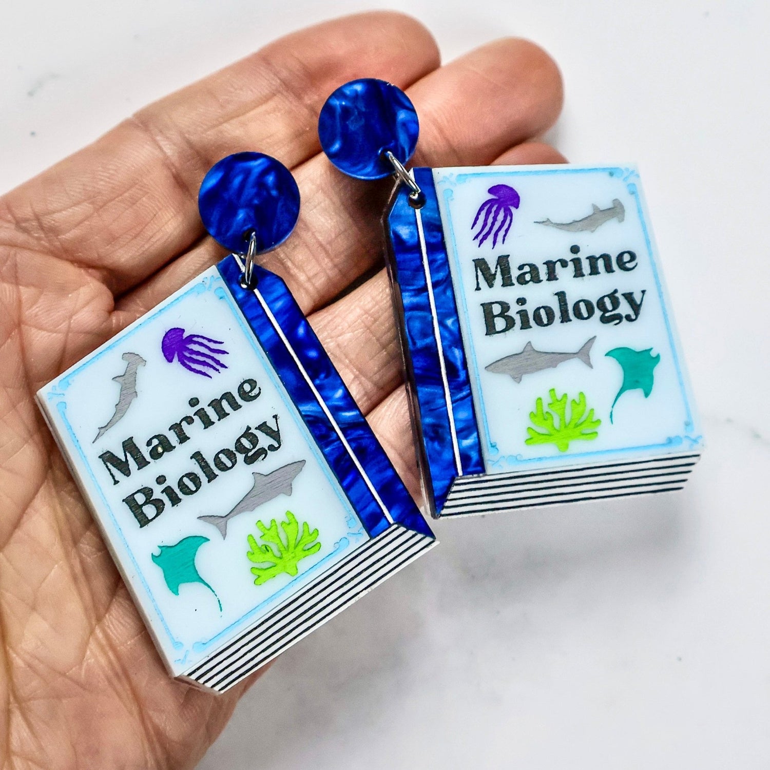 Earrings shaped like miniature books titled 'Marine Biology' held in a hand.