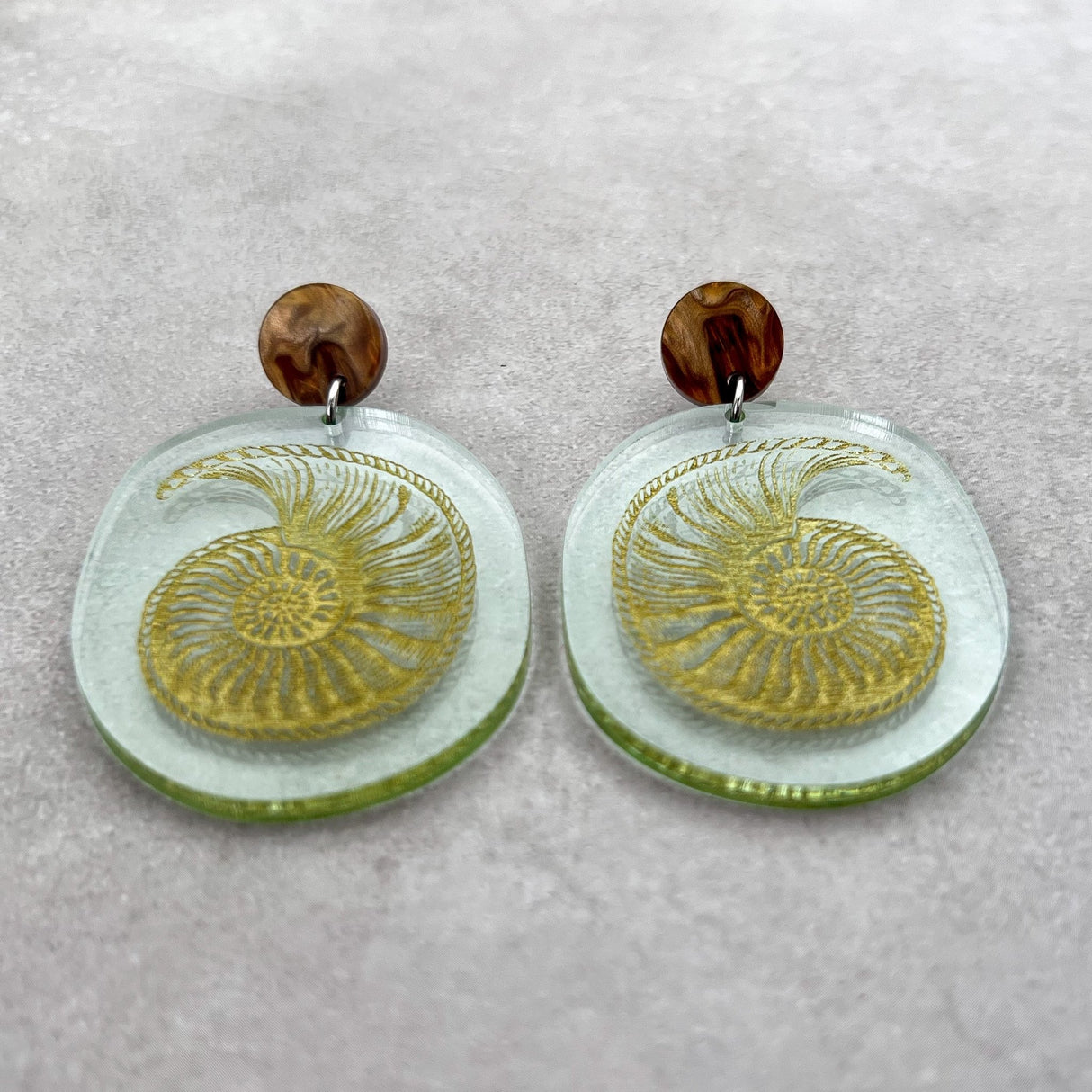 Nautilus fossil earrings, with a gold nautilus design handpainted on a green seaglass toned acrylic. They earrings hang from brown acrylic toppers. Front view.