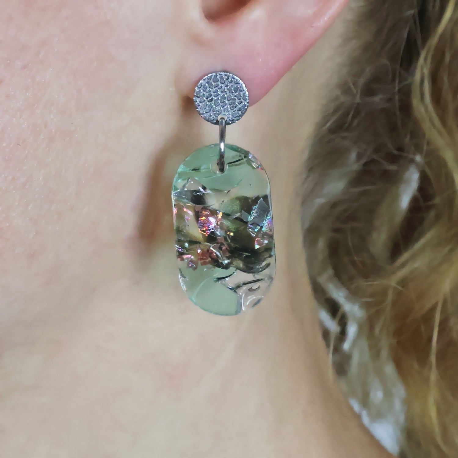 A pair of oval shaped recycled acrylic earrings with stainless steel toppers being modelled on an ear.