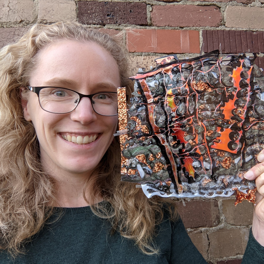 Louise, the maker, a woman with blonde hair and glasses, smiling and holding up a sheet of melt pressed acrylic.