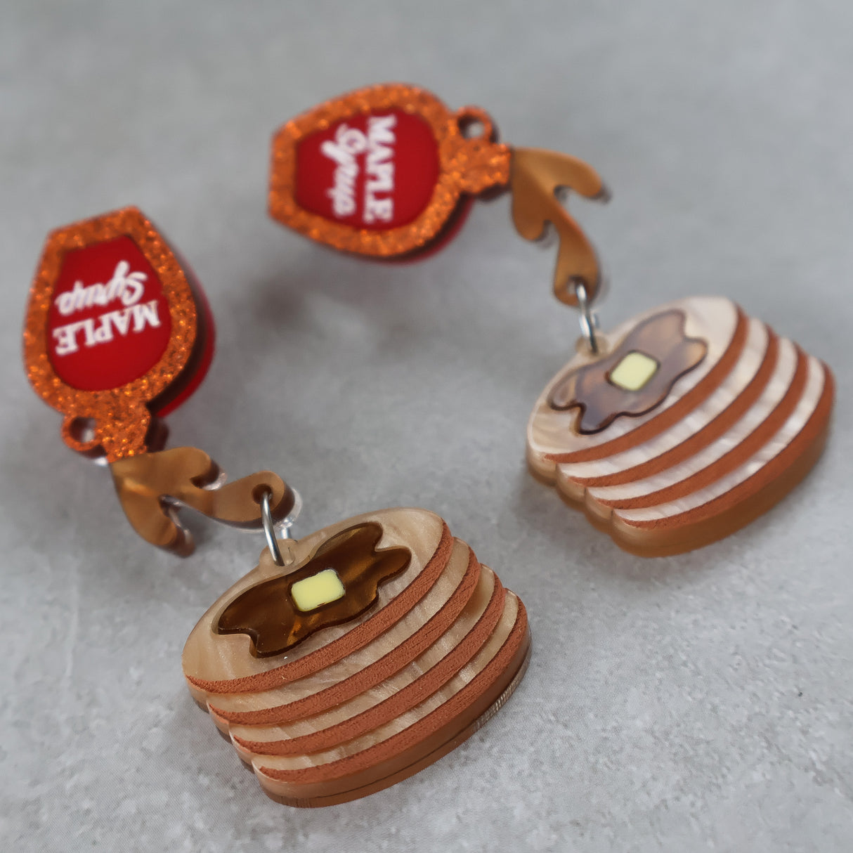 Acrylic earrings shaped like pancakes with syrup and butter on top, on a gray background.