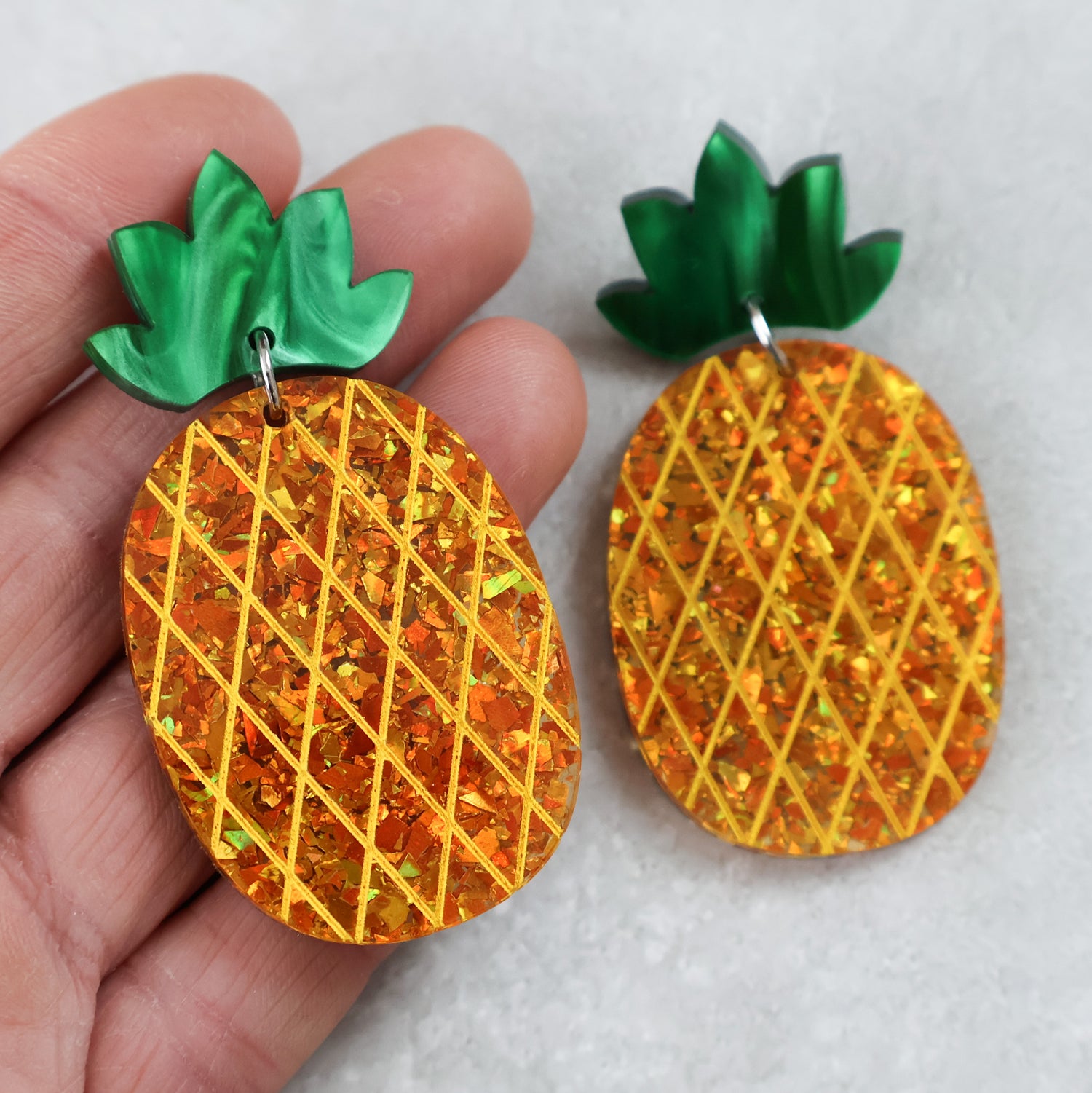 Pineapple-shaped earrings with gold glitter acrylic and green tops held by a hand on a light grey background.