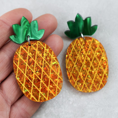 Pineapple-shaped earrings with gold glitter acrylic and green tops held by a hand on a light grey background.
