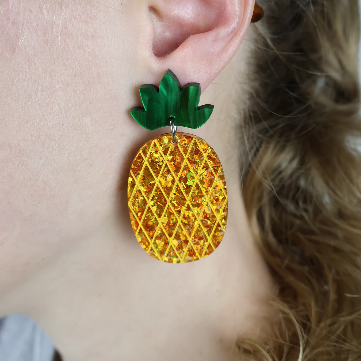 Gold glittery pineapple-shaped acrylic earrings with green toppers worn by a person with blonde hair.