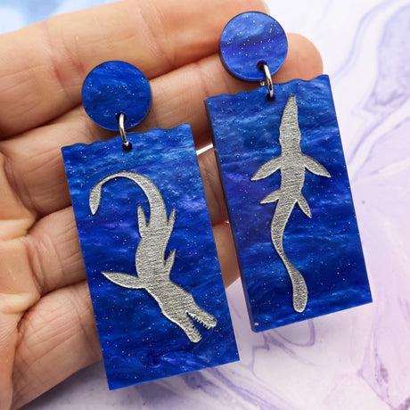A pair of asymmetrical blue acrylic dangle earrings featuring two different silver painted plesiosaurs being held in an hand against a marbled background.