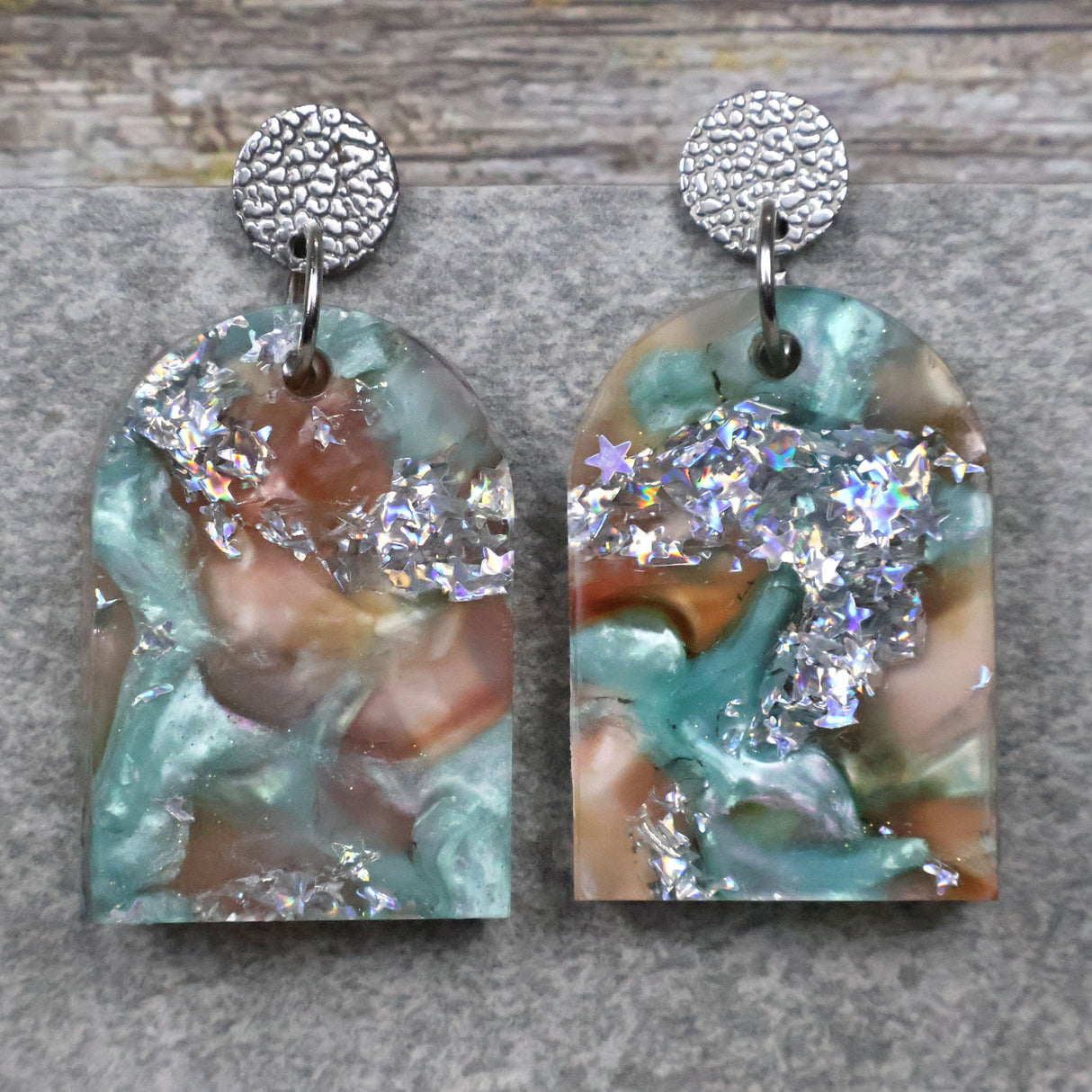A pair of one of a kind arch shaped recycled acrylic earrings with silver, green and brown design titled 'Celestial Tide' and stainless steel toppers. Pair 102-03