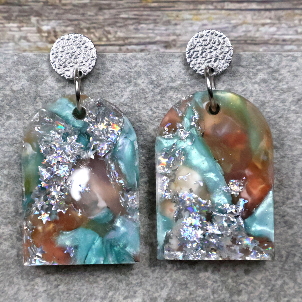 A pair of one of a kind arch shaped recycled acrylic earrings with silver, green and brown design titled 'Celestial Tide' and stainless steel toppers. Pair 102-06