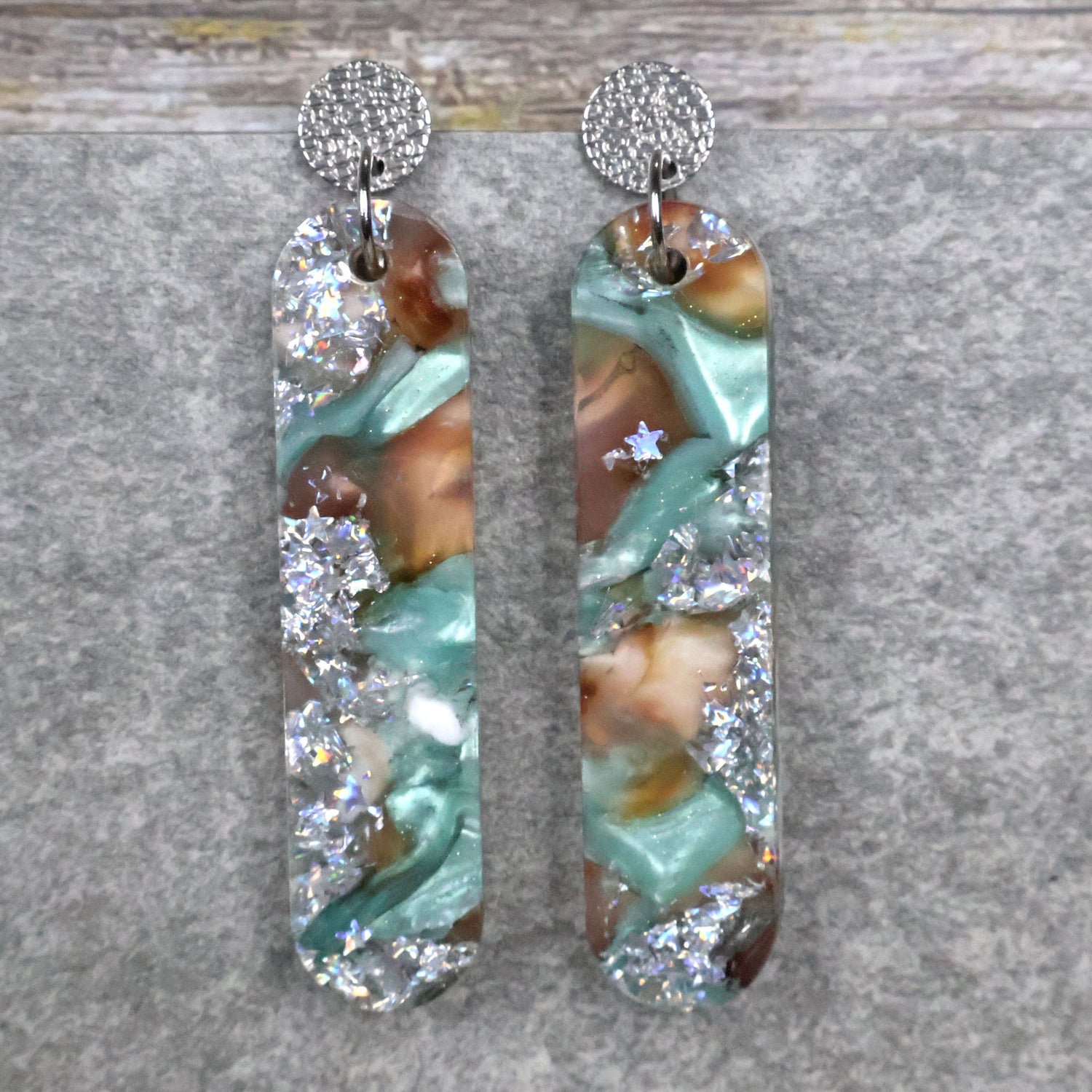 A pair of one of a kind long oval shaped recycled acrylic earrings with silver, green and brown design titled 'Celestial Tide' and stainless steel toppers. Pair 102-13
