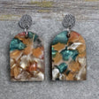 Pair of one of a kind arch shaped acrylic earrings with multicolored  design titled 'Sunset Reef' and stainless steel toppers on a gray background. Pair 106-04