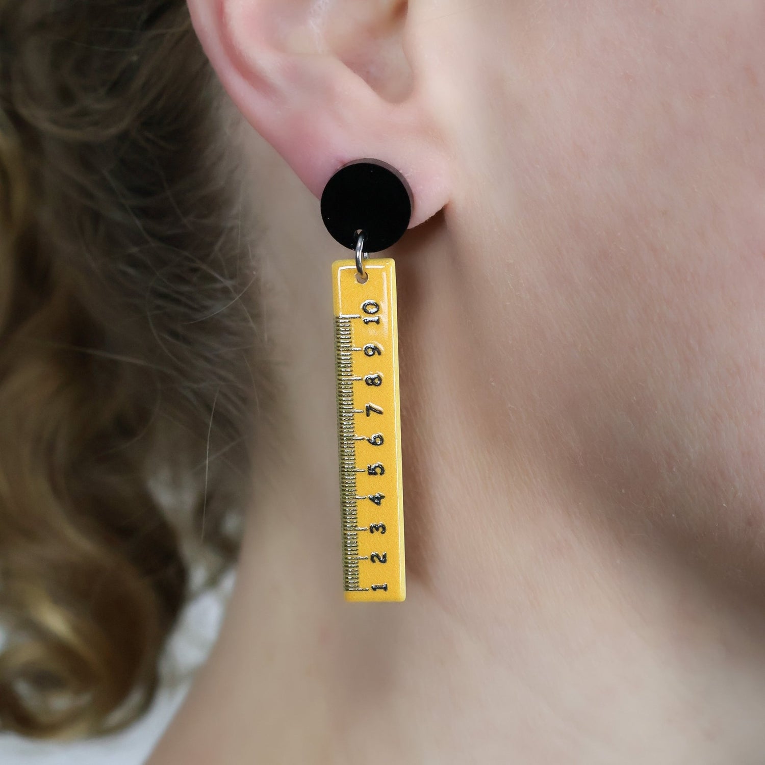 Yellow measuring ruler dangle earrings, hanging from black round acrylic toppers. Being modelled.