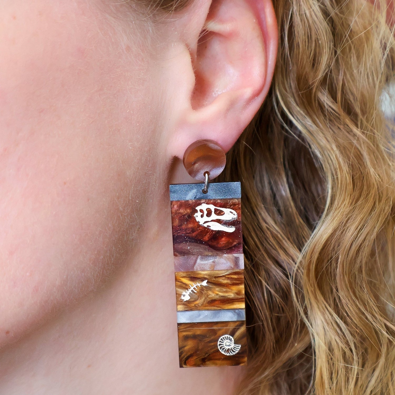 Dangle earrings representing sedimentary layers, comprising 6 strips of different grey and brown acrylics, with a t-rex skull, fish bones and nautilus engraved and handpainted in the larger layers. They hang from brown toppers and are being modelled in this photo.