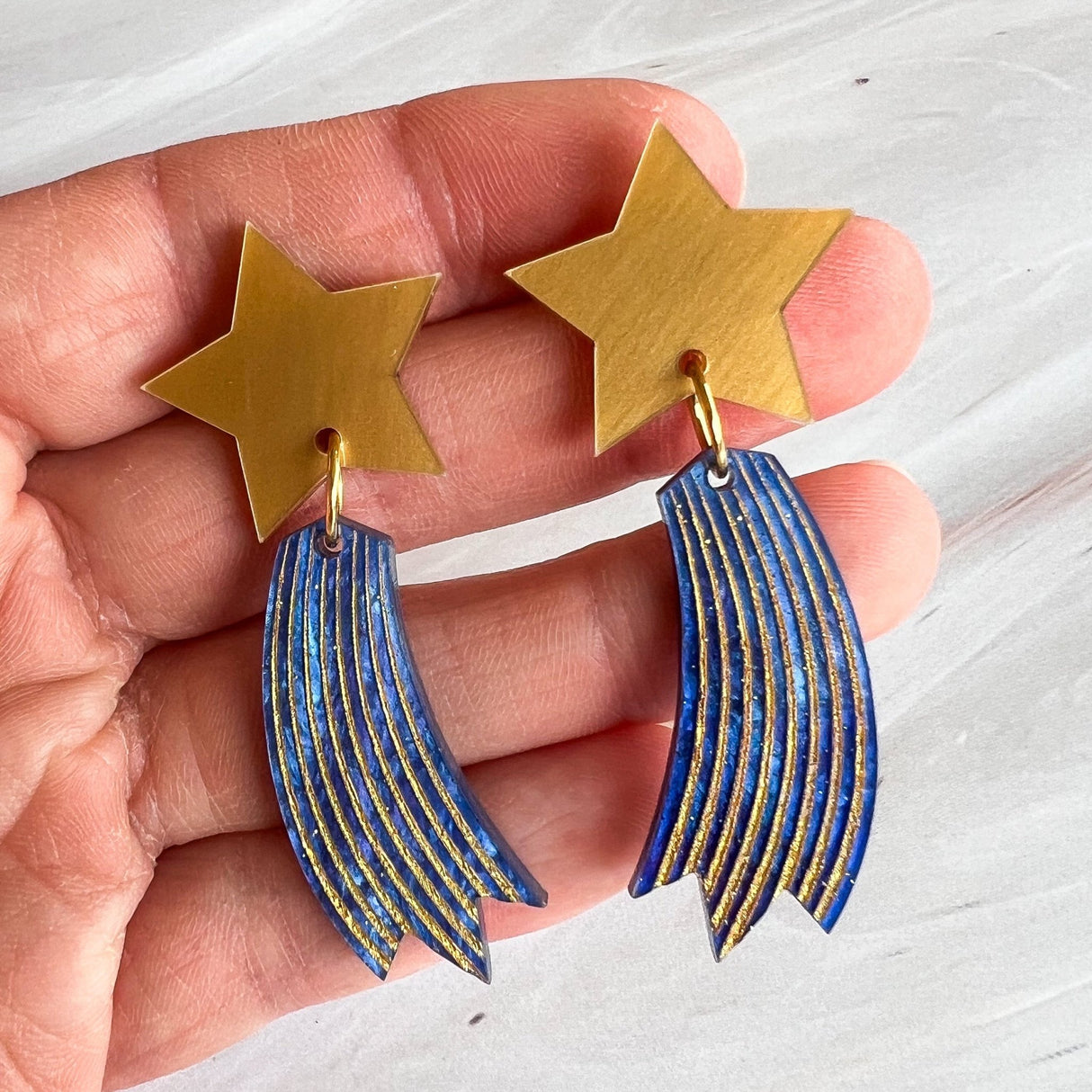 Shooting star earrings, comprised of gold toned acrylic star toppers with blue and gold star trails dangling underneath. Earrings are being held.