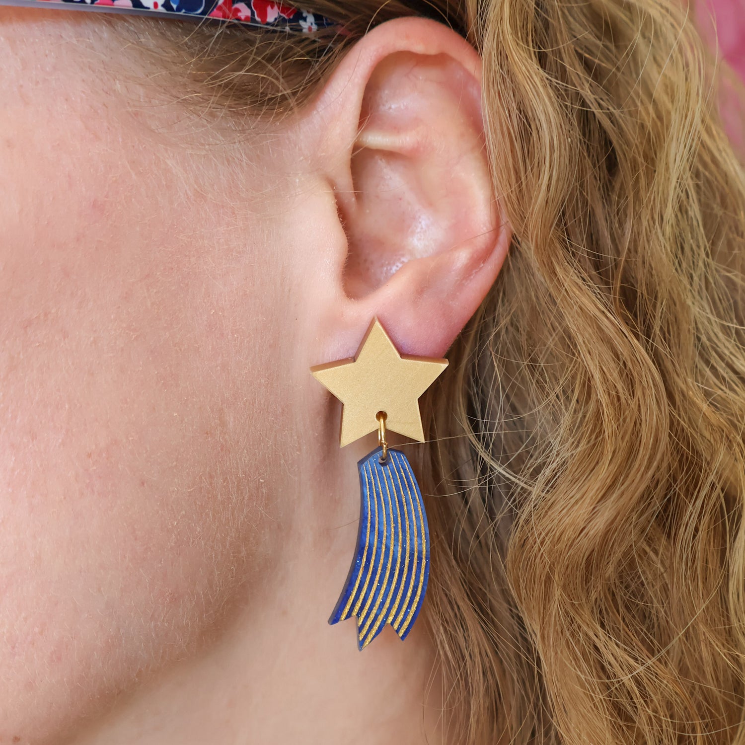Shooting star earrings, comprised of gold toned acrylic star toppers with blue and gold star trails dangling underneath. Earrings are being modelled.
