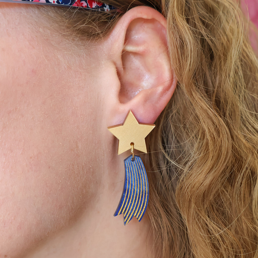 Shooting star earrings, comprised of gold toned acrylic star toppers with blue and gold star trails dangling underneath. Earrings are being modelled. 