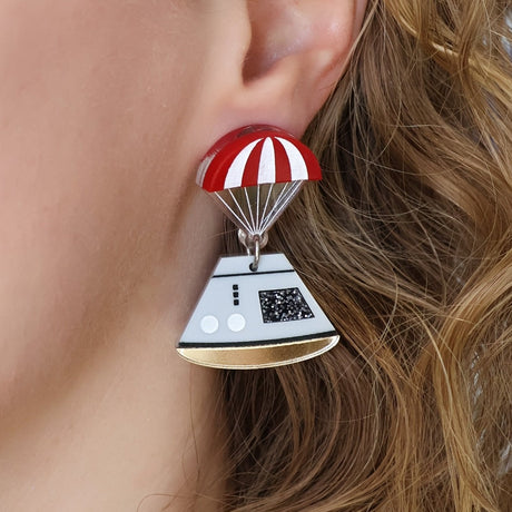 Space capsule earrings, with and red parachute topper and a grey capsule dangling underneath. Laser cut from acrylic. Earrings are being modelled.