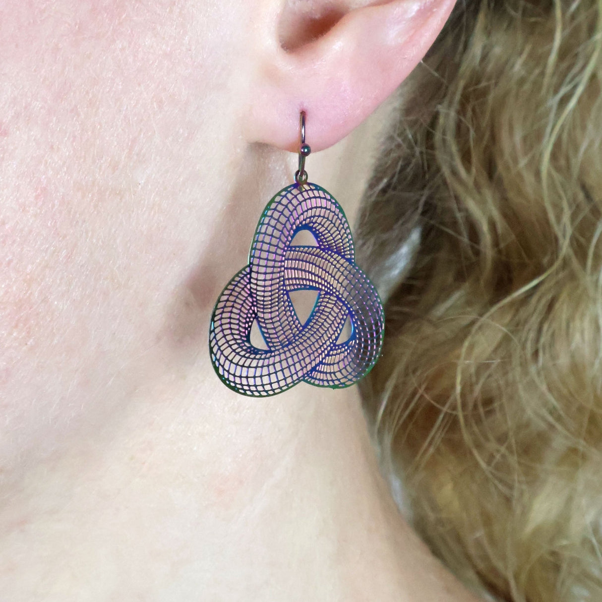 Close-up of a person wearing geometric surface themed earrings in a a rainbow toned stainless steel with french hook tops.
