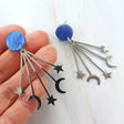 Earrings with blue round toppers and star and moon stainless steel dangles being held in a hand against a white background.