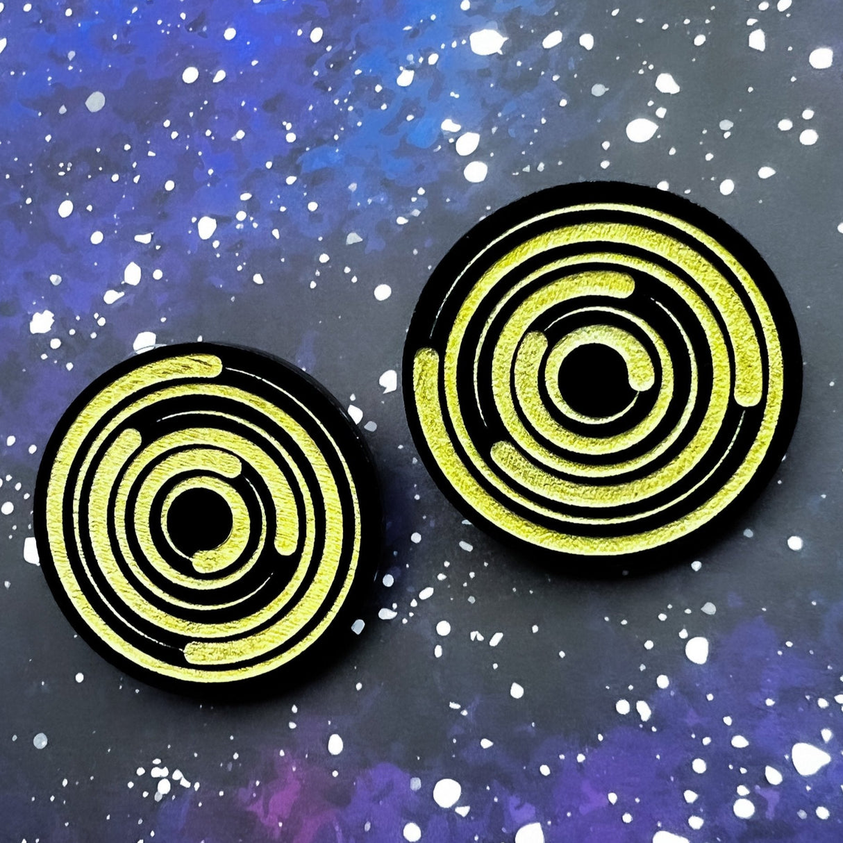 A pair of round acrylic star trail studs, black with gold painted star trails within.