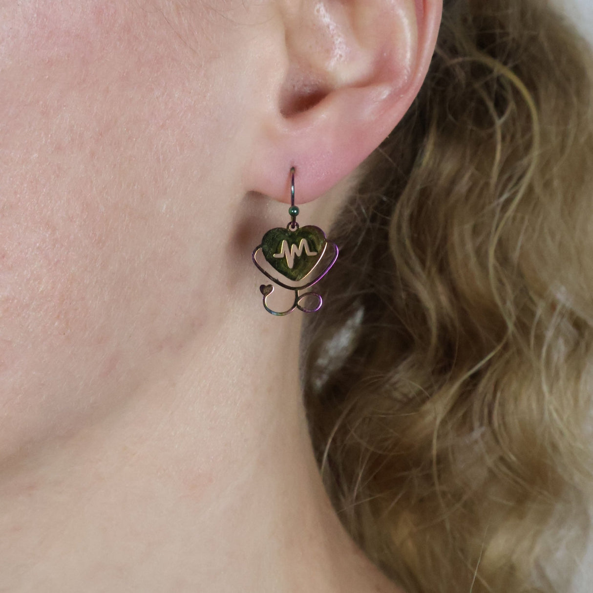 Small stethoscope themed earrings a heart and heartbeat design in rainbow stainless steel worn by a person with blonde hair.