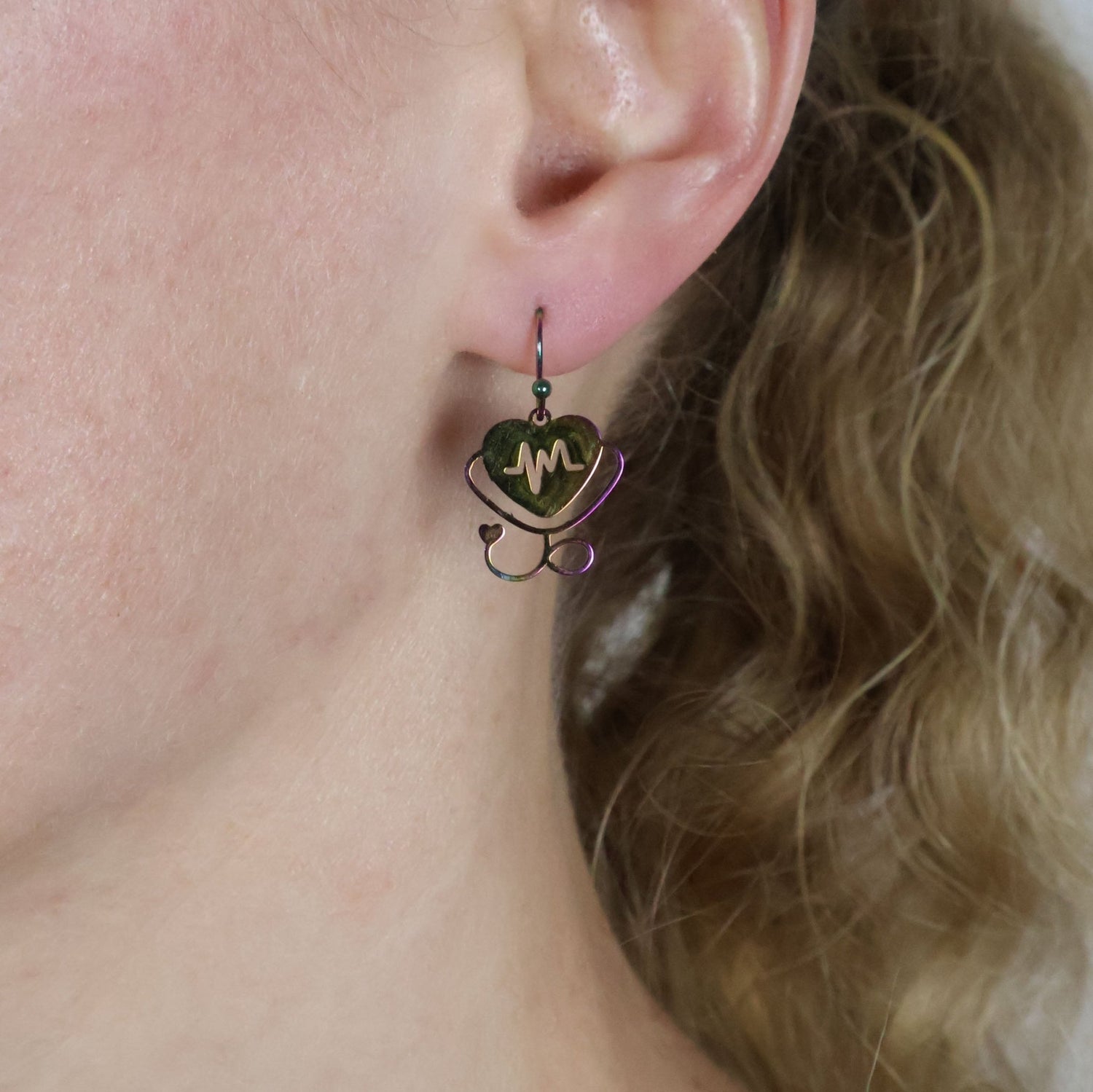 Small stethoscope themed earrings a heart and heartbeat design in rainbow stainless steel worn by a person with blonde hair.