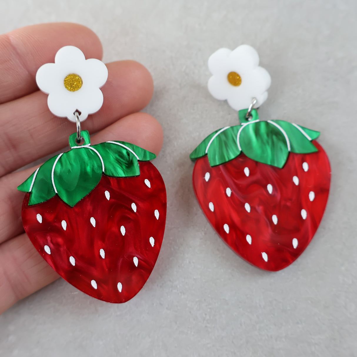 Strawberry-shaped marbled acrylic earrings with white flower toppers held by a hand on a light grey background