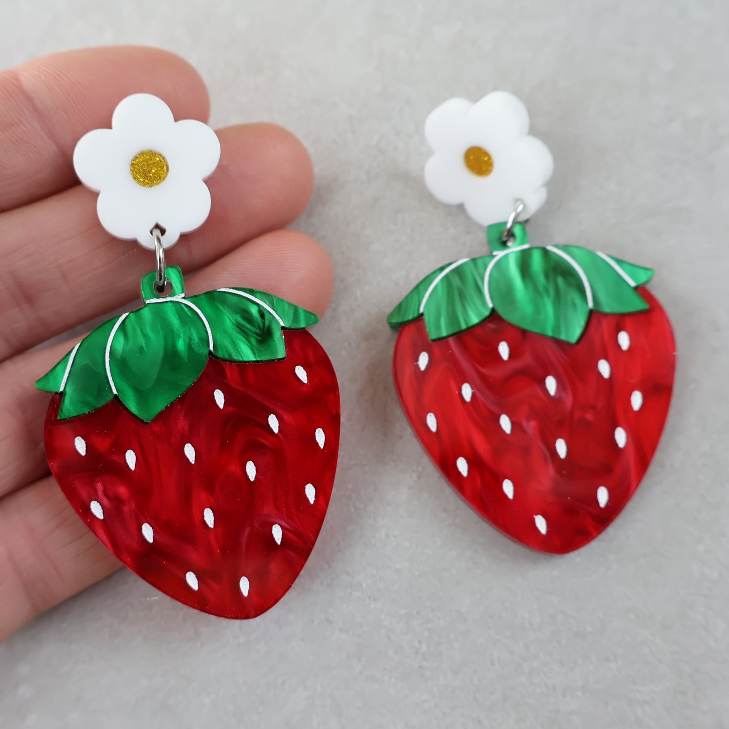 Strawberry-shaped marbled acrylic earrings with white flower toppers held by a hand on a light grey background