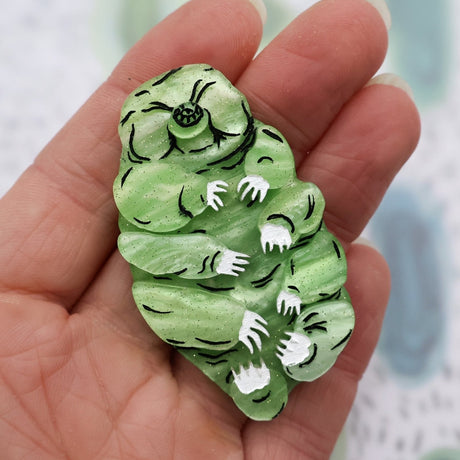 Green acrylic laser cut tardigrade brooch in hand for scale. Brooch has black and white handpainted details.