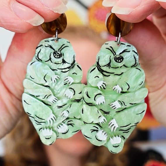 Tardigrade earrings made from swirly apple green acrylic, hanging from swirly brown round earring toppers. Earrings are being held up, and there is an out of focus person in the background. 