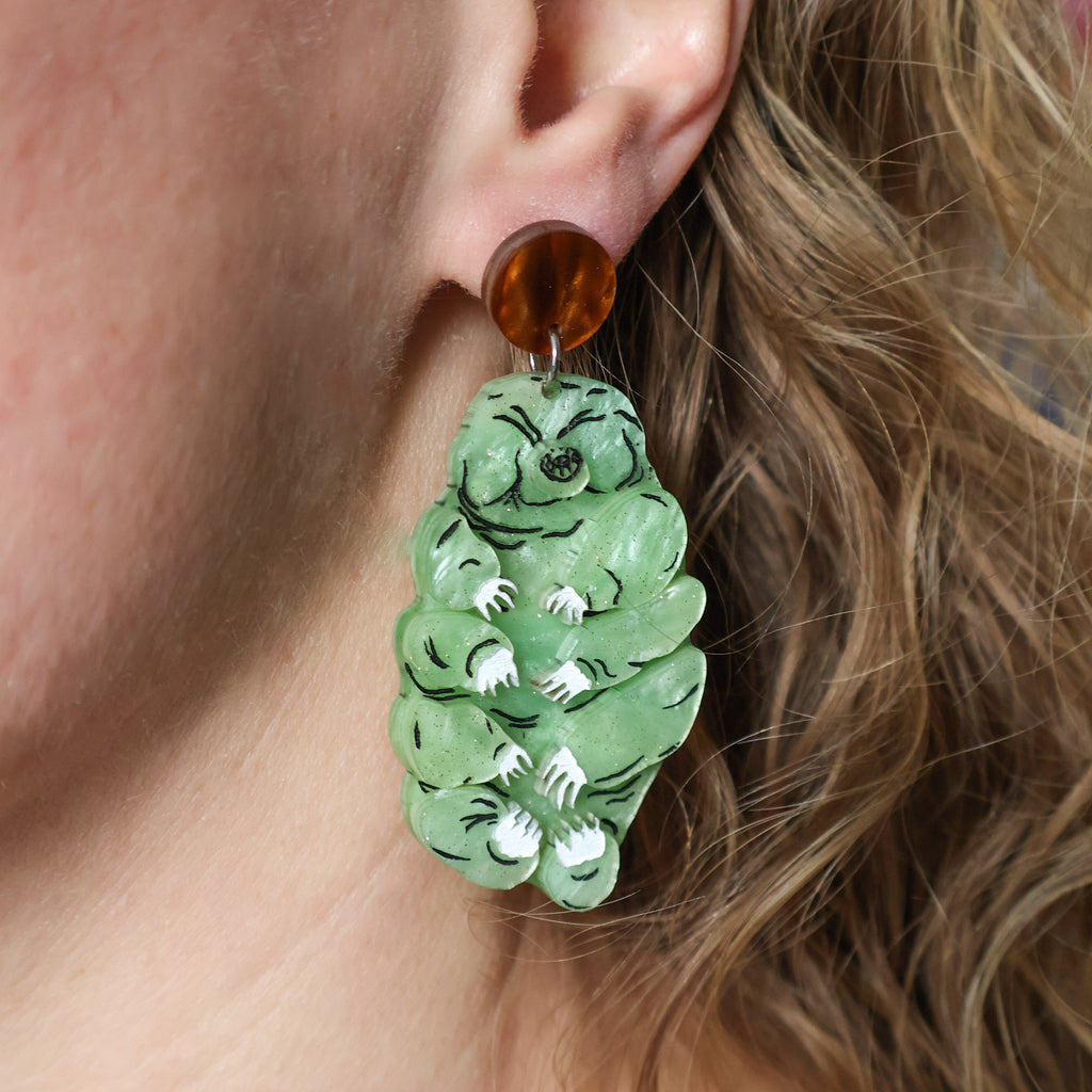 Tardigrade earrings made from swirly apple green acrylic, hanging from swirly brown round earring toppers. Earrings are being modelled.  
