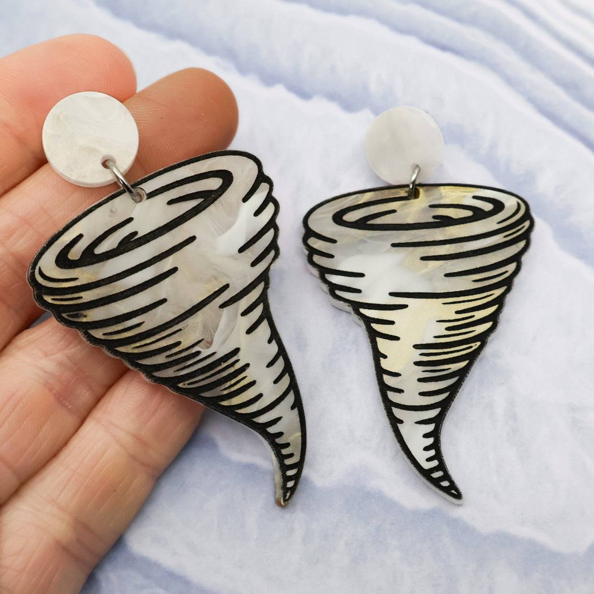 Tornado-shaped acrylic earrings held between fingers on a light blue background