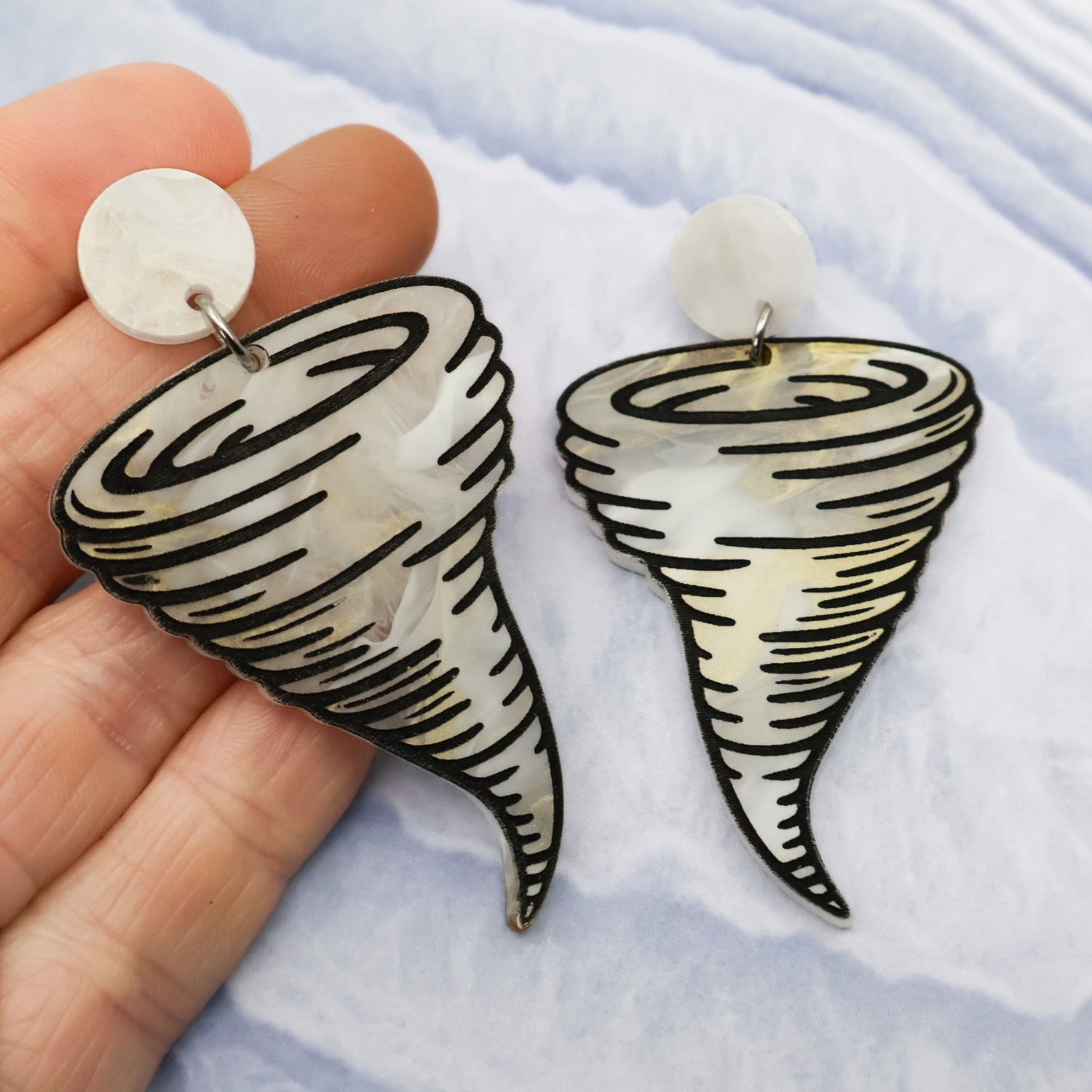 Tornado-shaped acrylic earrings held between fingers on a light blue background